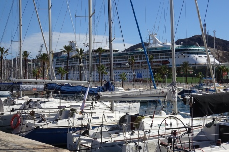 Large sailboats in the yacht harbor line the foreground. The ship is behind with a mountain in the far distance.