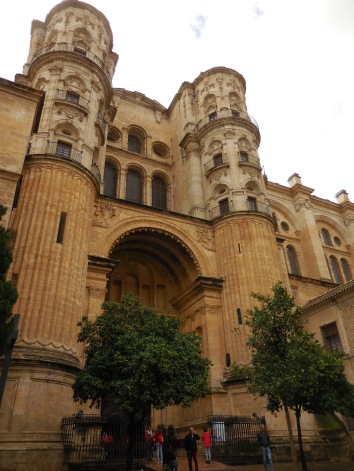 Close up of the Renaissance style cathedral. People are standing outside a huge door. The stone facade has sandstone columns beside the door and square stones above. The top is lined with stained glass windows.