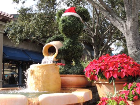 A very large topiary of Mickey Mouse pouring water into a large jug with water then cascading down other containers. He is wearing a red Santa hat with a white, fur band. There are planters of poinsettias beside and a tree, still with leaves, behind.