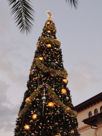 The top portion of a large Christmas tree rising above the rooftops. It has massive ornaments lit with Edison light bulbs. At the top of the photo, in the foreground are two palm fronds from a nearby tree.