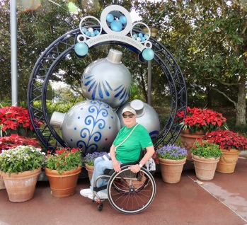 George is sitting in front of a display of large tree ornaments and is surrounded by rotted poinsettias and other blooming flowers. He is wearing a green shirt, jeans, white Converse shoes, and a tan visor.