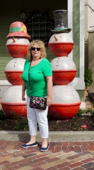 Sandy is standing between, slightly to the front of, 2 snowmen. Each is made of white and red balls. The female has a red hat, and the male has a black top hat. Sandy is wearing a green top, white jeans, and blue skimmers.