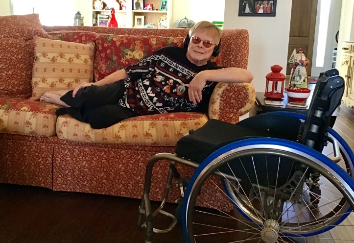 George is stretched out on the sofa. He has blond hair and glasses with an orangish tint. He is wearing a black, Mickey and friends sweater with white snowflakes, black pants, black headphones, and he is barefooted. 