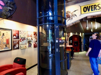 In the center is the elevator. It has a blue, steel frame with a clear tube and a door. It is just large enough to carry a wheelchair. To the right is a young man with glasses, a navy shirt, and jeans. On the left are posters and pictures on the wall.