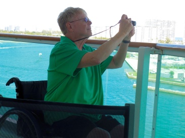 George is sitting on the balcony of a cruise ship taking a photo with a small camera. He has rimless glasses with a gray tint. He is wearing a bright green polo shirt.