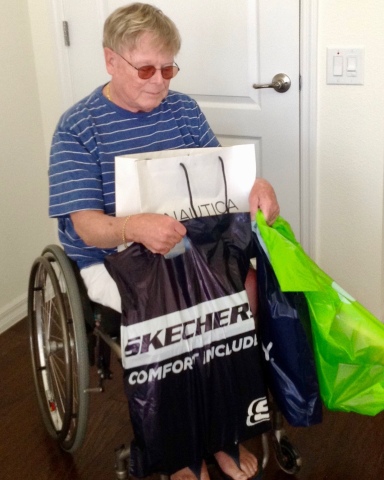George is in his wheelchair, at home, with shopping bags from Skechers, Nautica and a brilliant green bag. He has blond hair, wire rimmed glasses with an orangish tine, a blue T-shirt with white stripes, and he has removed his shoes.