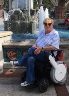 George is sitting in a state-of-the-art power wheelchair. He is in front of a large fountain. He has blonde hair, a striped shirt, jeans, and blue Converse shoes. One leg is crossed over the other.