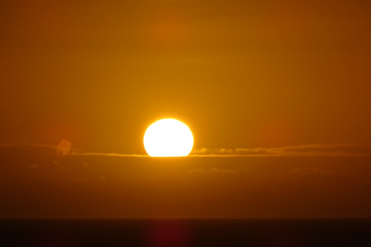 A hot, hazy sunset. The white sun is setting below a cloud bank is an orange sky.